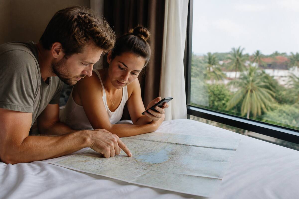 Een stel dat vakanties en reisroutes vergelijkt met documenten en een telefoon, bij een hotelraam met uitzicht op palmbomen.
